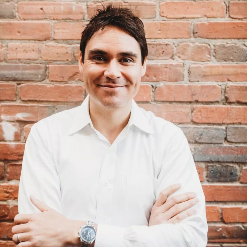 The image shows a man with dark hair and a white shirt posing in front of a brick wall. He has a watch on his left wrist and his arms are crossed. He is smiling at the camera. The background is a blurred brick wall.